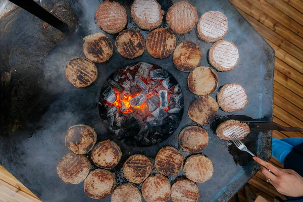 Hamburger köfteleri parlayan kömürün üstünde çatal ve spatula kullanarak mangalda cızırdıyor. Buhar ve ızgara izleri görünür..