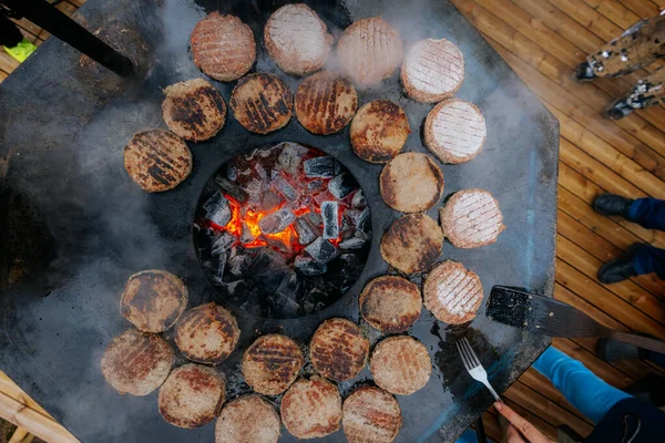 Hamburger köfteleri parlayan kömürün üstünde çatal ve spatula kullanarak mangalda cızırdıyor. Buhar ve ızgara izleri görünür..