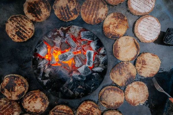 Hamburger köfteleri parlayan kömürün üstünde çatal ve spatula kullanarak mangalda cızırdıyor. Buhar ve ızgara izleri görünür..