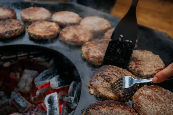 Hamburger köfteleri parlayan kömürün üstünde çatal ve spatula kullanarak mangalda cızırdıyor. Buhar ve ızgara izleri görünür..
