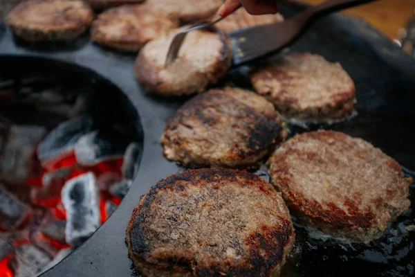 Hamburger köfteleri parlayan kömürün üstünde çatal ve spatula kullanarak mangalda cızırdıyor. Buhar ve ızgara izleri görünür..