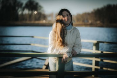 Beyaz kışlık ceketli genç bir kadın gölün kenarındaki ahşap korkuluklara yaslanmış, sakin bir ifadeyle yukarıya bakıyor. Ilık doğal ışık yumuşak, rüya gibi bir atmosfer yaratır..