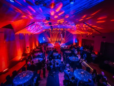 Yuvarlak masalarla dolu büyük bir ziyafet salonu ve canlı kırmızı, mavi ve mor ışıklar ve yansımalarla aydınlatılmış resmi giyinmiş konuklar. 11.19. 2025 Valmiera, Letonya