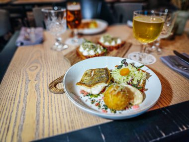 A plated gourmet fish dish with crispy skin, served alongside a salad and fried ball, placed on a wooden table with beer and other appetizers in a restaurant setting.