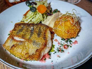 A plated gourmet fish dish with crispy skin, served alongside a salad and fried ball, placed on a wooden table with beer and other appetizers in a restaurant setting.