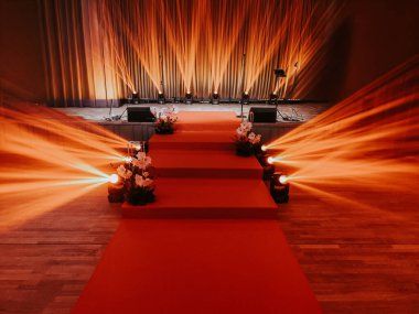 A brightly lit stage with a red carpet, floral decorations, and orange spotlights leading up to a curtained backdrop and microphones, prepared for a formal event or performance.