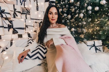 Beyaz süveter içinde şık bir kadın figürü, sarılı hediyeler ve güzel ışıklandırılmış bir Noel ağacı ile çevrili, rahat bir tatil havası yayıyor.