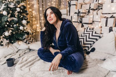 Mavi pijamalı bir kadın, Noel ağacı ve hediyelerle çevrili lüks bir halının üzerinde oturuyor. Yumuşak dokuları ve ılık ışıklandırmalarıyla rahat bir tatil havasını çağrıştırıyor.