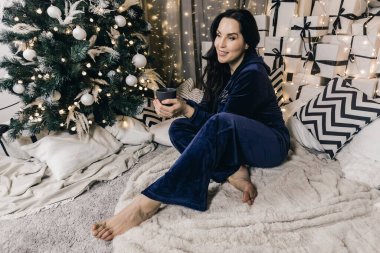 Mavi pijamalı bir kadın yumuşak bir battaniyede gevşiyor, elinde içki, arka planda Noel ağacı ve dekoratif hediyeler var, sıcak bir tatil ortamı yaratıyor.