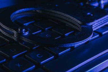 Bir klavyeye yerleştirilen kelepçeler, mavi ışıkla vurgulanmış, suç, adalet temaları ve teknoloji ile kolluk kuvvetlerinin kesişme noktasını temsil ediyor.