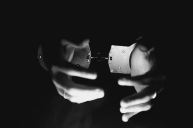 Kişinin elleri kelepçeleri kavrıyor, hafif ışıklandırma ile vurgulanıyor, karanlık bir zemin üzerine kuruluyor, kısıtlama ve kontrol temaları çağrıştırıyor