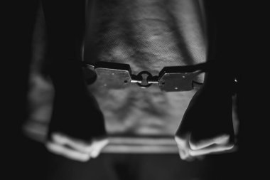 Elleri kelepçeli, metal bir çubuğu kavrayan, karanlık bir zemin üzerine kurulmuş, adalet temaları, hapsetme, ve hukuk sistemini çarpıcı bir monokrom görüntüsüyle betimleyen bir birey.