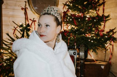 Beyaz kürklü kadın ve taç, sıcak bir ortamda oturmuş, güzel dekore edilmiş bir Noel ağacı, zarafet ve tatil ruhu yayıyor.