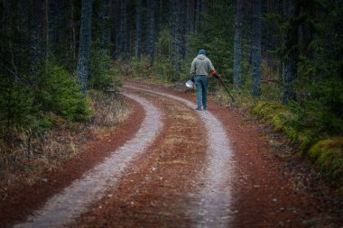 Ormanda dolambaçlı bir yolda yürüyen kişi, elinde bakım araçları, ağaçlar ve toprak renklerle çevrili, açık hava keşfi ve doğanın özünü yakalıyor.