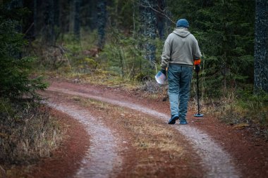 Metal dedektörlü bir birey, bereketli bir orman boyunca, etrafını ağaçlar ve çalılıklar sarmış, doganın huzurunun tadını çıkararak dolambaçlı toprak bir yolda yürüyor.