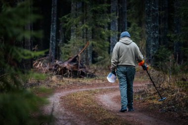 Gri kapüşonlu ve mavi pantolonlu kişi ağaçlar ve yapraklar arasında metal dedektör kullanarak orman yolunda yürüyor, açık havada keşif ve keşif yapıyor.
