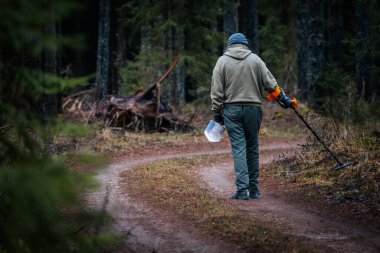 Çevresi yemyeşil ve toprak dokularla çevrili bir orman yolu boyunca metal detektörüyle uğraşan kişi, açık hava hazine avcılığı macerasını vurguluyor.