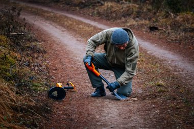 Erkek hazine avcısı, doğal elementlerle çevrili ormanlık bir alanda toprak bir patikayı kazıyor. Macera ve keşfin heyecanını vurguluyor.