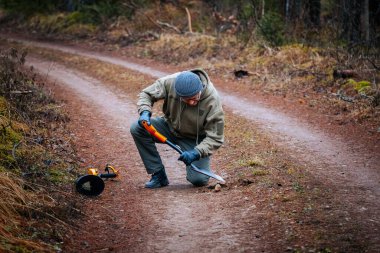 Açık hava meraklısı, ağaçlar ve toprak dokularla çevrili bir el bilgisayarı kullanarak bir orman yolunda diz çöküyor, doğa keşiflerini vurguluyor.
