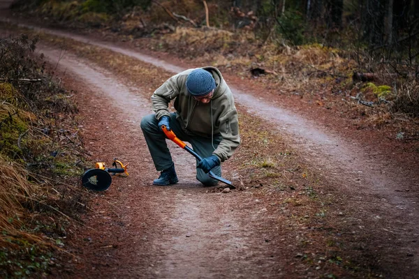 Açık hava meraklısı bir orman yolunda diz çöküyor, bir aletle toprağı kazıyor, ağaçlar ve doğal elementlerle çevrili, keşif ve macerayı vurguluyor.