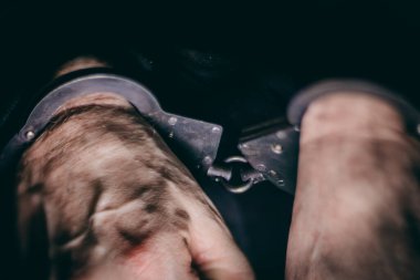 Erkek elleri metal kelepçelerle korunuyor, deri ve kelepçelerin dokusunu vurguluyor, kısıtlamanın ve adaletin güçlü bir görsel anlatımı yaratıyor.