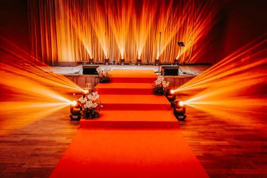 Kırmızı halı yolu, çiçek aranjmanlarıyla çevrili sıcak turuncu ışıklı bir sahneye çıkıyor. Toplantılar için davetkar ve şenlikli bir ortam yaratıyor.