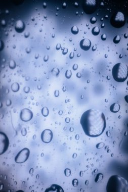 Camdaki su damlacıklarının yakın plan görüntüsü, yumuşak mavi tonlarda soyut bir desen sergiliyor, rahatlatıcı bir atmosfer yaratıyor ve doğanın güzelliğini ön plana çıkarıyor.