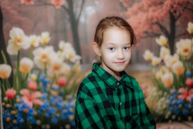 Açık kahverengi saçlı, yeşil ekose gömlekli çocuk renkli çiçek arkaplanına karşı poz veriyor. İlkbahar güzellikleriyle dolu neşeli ve canlı bir sahne yaratıyor.