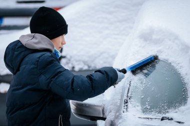 Kışın giydiği çocuk, karlı bir ortamla çevrili bir fırça kullanarak arabanın ön camındaki karı kaldırıyor ve kış havasının zorluklarını vurguluyor.