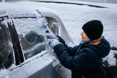 Kışın giydiği çocuk, karlı bir manzarayla çevrili bir arabanın ön camından kar kazıyarak kış aktivitelerinin ve açık hava oyunlarının neşesini somutlaştırıyor.