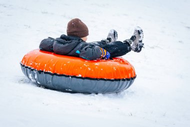 Valmiera, Latvia - January 02, 2026: Boy in black jacket and brown beanie is snow tubing on orange inflatable tube in snowy environment