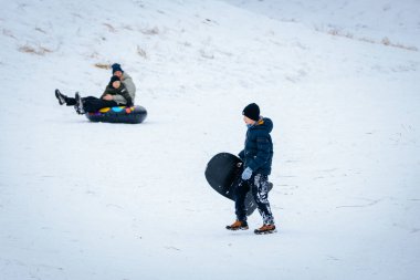 Valmiera, Letonya - 02 Ocak 2026: Mavi ceketli çocuk kızakla karlı tepede yürürken, başka bir çocuk renkli şişme tüpte kayıyor