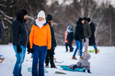 Valmiera, Letonya - 2 Ocak 2026: Turuncu ceketli kadın, karlı tepedeki adamla birlikte, aileler arka planda kış etkinliklerinin keyfini çıkarıyor
