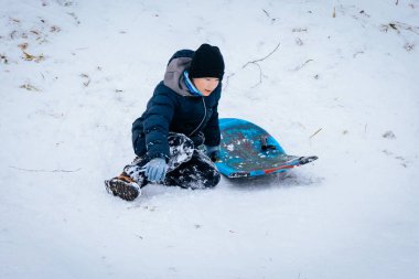 Child in winter attire sits in snow next to a vibrant sled, surrounded by a snowy landscape, capturing the joy of outdoor winter activities and playfulness