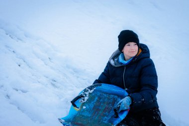Kızaklı genç adam, ılık kış kıyafetleri içinde, karlı tepede oturuyor, mutlu bir şekilde gülümsüyor, dingin bir kış manzarasıyla çevrili, taze karla kaplı.