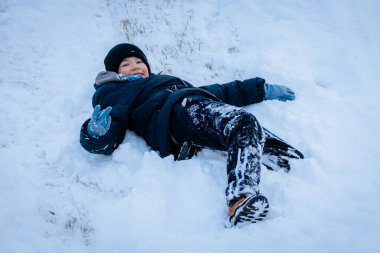 Kışın giyinmiş çocuk mutlu bir şekilde karda uzanıyor, soğuk havanın tadını çıkarıyor, neşeli bir ifadeyle ve karla kaplı çevresiyle neşeli bir sahne yaratıyor.