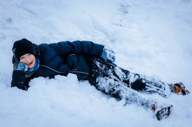 Kışın giydiği çocuk, kar taneleriyle dolu sakin bir kış ortamı ile çevrili, yumuşak karda uzanıyor, gülümsüyor ve soğuk havanın tadını çıkarıyor.