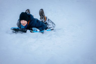 Mutlu genç çocuk kar kaplı bir tepede kayıyor, mavi bir ceket ve siyah bir şapka giyiyor, kış mevsiminin tadını çıkarıyor, karla kaplı manzarası ve neşeli bir atmosferi var.