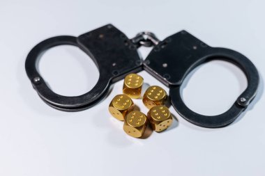 Kelepçeler altın zarların yanında beyaz bir yüzeyde sergileniyor, eşsiz ve düşündürücü bir düzende şans ve riskin görsel bir anlatımını oluşturuyor.