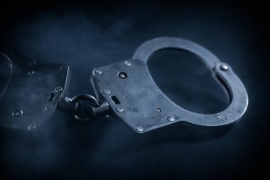 Metalik kelepçeler karanlık bir yüzeye yerleştirilmiş, dumanlı bir atmosferle kaplanmış, kanun uygulama, adalet ve dizginleme temalarını temsil ediyor.