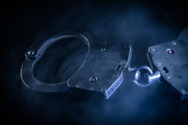 Metalik kelepçeler karanlık bir yüzeye yerleştirilip sisle kaplanarak adalet ve kanun uygulama temalarını çağrıştıran karamsar bir atmosfer yaratıyor.