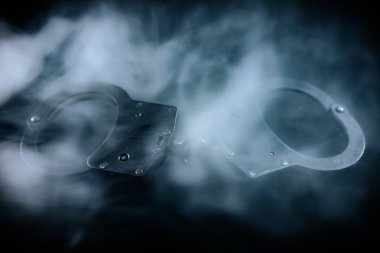 Karanlık bir yüzeye yerleştirilmiş kelepçeler, dumanla kaplanmış, suç, adalet ve entrika temalarını ortaya koyan karamsar bir ortam yaratıyor.