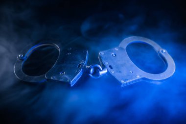 Mavi dumanla çevrili, karanlık bir yüzeye yerleştirilmiş kelepçeler kolluk kuvvetleri ve güvenlikle ilgili esrarengiz ve yoğun bir ortam yaratıyor.