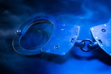 Kelepçeler karanlık bir yüzeyde sergileniyor. Mavi bir sisle örtülmüş. Adalet, kolluk kuvvetleri ve suçun ve cezanın karmaşık doğasını temsil ediyor.