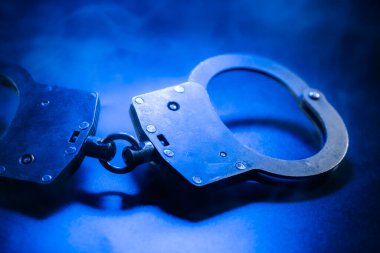 Metalik kelepçeler, mavi ışıkla vurgulanmış karanlık bir yüzeye yerleştiriliyor. Kanun uygulama ve adalet temalarını çağrıştıran dengesiz bir ortam yaratıyor.