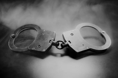 Karanlık bir yüzeye kelepçelenmiş, dumanla kaplanmış, kolluk kuvvetleri, adalet ve kısıtlama temaları çarpıcı bir siyah beyaz kompozisyonda temsil ediliyor.