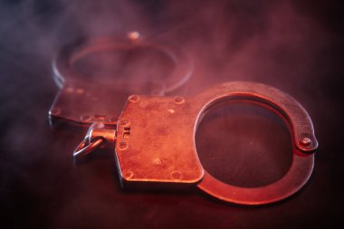 Karanlık yüzeyde metalik kelepçelerin yakın plan çekimleri, dumanla kaplanmış, kanun ve adalet temalarını çağrıştıran dengesiz bir ortam yaratıyor.