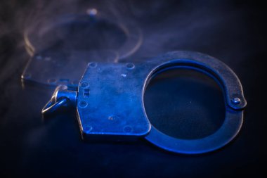 Kelepçeler karanlık yüzeyde, mavi ışıkla vurgulanmış, kasvetli bir ortam yaratıyor, kanun ve adalet temalarını temsil ediyor.