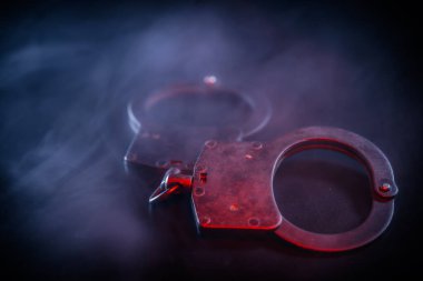 Metalik kelepçeler karanlık bir yüzeye yerleştirilmiş, dönen dumanla kaplanmış, kolluk kuvvetleri, adalet ve olay yerindeki gerilimi çağrıştırıyor.