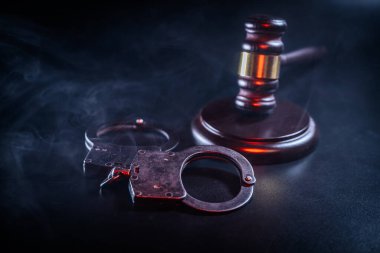 Karanlık bir yüzeyde bir tokmağın yanına yerleştirilmiş kelepçeler, dumanlı bir ortam, adalet temaları, kolluk kuvvetleri ve hukuk sistemi temaları.